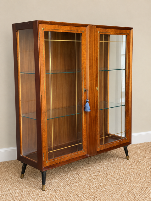 Angled view of a vintage wooden display cabinet showing its glazed side panel, two glass doors with gold detailing, glass shelves and tapered black legs